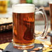 A clear glass 12 oz. beer mug filled with amber beer and topped with foam, placed on a napkin with assorted nuts nearby.