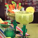 A 12 oz. margarita glass with a green cactus-shaped stem, shown filled with a margarita and garnished with a lemon slice.