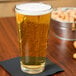 A clear, rim-tempered stackable mixing glass filled with a light, carbonated beverage, placed on a dark napkin on a wooden table.