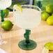 A 16 oz. margarita glass with a green cactus-shaped stem, filled with a margarita and garnished with a lime wedge.