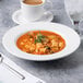 A white porcelain soup bowl filled with vegetable soup and garnished with a sprig of rosemary.