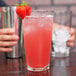 A clear 16 oz. tall mixing glass filled with a pink iced beverage and garnished with a strawberry on the rim.