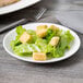 A small white porcelain coupe plate holding a simple salad with lettuce and croutons.