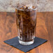 A clear 16 oz. cooler glass filled with a dark carbonated beverage and ice, placed on a black napkin on a wooden surface.