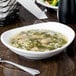 A bright white oval porcelain bowl filled with soup, placed on a wooden table.