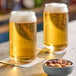 A clear 16 oz. can-shaped glass filled with beer, placed on a coaster next to a small bowl of mixed nuts.