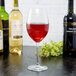 A clear 17 oz. wine glass filled with red wine, surrounded by bottles of wine on a dark surface.