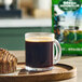 A clear glass mug of black coffee sits on a wooden tray next to a chocolate muffin, with a bag of Green Mountain Coffee Roasters Sumatra Reserve Whole Bean Coffee in the background.