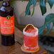 A bottle of Liber & Co Blood Orange Cordial Syrup next to a glass of red cocktail with a sugared rim and a dried orange garnish.