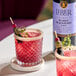 A glass of pink cocktail garnished with berries and herbs, next to a bottle of Liber & Co Marion Blackberry Syrup.