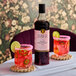 A bottle of Liber & Co Pacific Strawberry Syrup with two glasses of a pink beverage garnished with lime slices and pink sugar rims.
