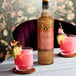 A 750 mL bottle of Liber & Co Almond Orgeat Syrup displayed on a table with two pink cocktails garnished with fruit.