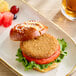 A plant-based vegan chik patty sandwich with lettuce and tomato on a pretzel bun, served with a side of fruit.