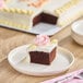 A slice of chocolate cake with white buttercreme icing and decorative pink and yellow frosting on a plate, with the remaining sheet cake in the background.