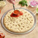 A 12-inch par-baked pizza crust on a metal tray with pizza sauce being spread on top.