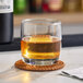 A clear Acopa Convex 10 oz. rocks glass filled with a brown beverage and a large ice cube, placed on a woven coaster.