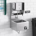 A stainless steel hands-free hand sink with a knee-operated valve, paper towel dispenser, and soap dispenser mounted on a tiled wall.