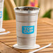 A 20 oz. aluminum cup with a blue 'Top Cup' logo, filled with ice and a dark beverage, placed on a wooden table.