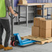 A blue manual pallet jack with 16-inch by 36-inch forks being used to move a wooden pallet loaded with cardboard boxes in a warehouse setting.