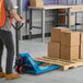 A blue manual pallet jack being used to move a wooden pallet loaded with cardboard boxes in a warehouse setting.