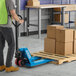 A blue manual pallet jack with 21-inch by 48-inch forks being used to move a wooden pallet loaded with cardboard boxes in a warehouse setting.