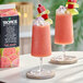 A carton of Tropics Mixology Pink Guava Frozen Beverage Mix next to two glasses of pink guava frozen drinks garnished with fruit and whipped topping.