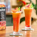 A pair of tall glasses filled with a peach-colored frozen beverage, garnished with peach slices and mint, with a carton of Tropics Mixology Peach Frozen Beverage Mix in the background.