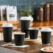 A set of black double wall ripple paper hot cups with white lids, displayed on a wooden surface.