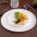 An ivory round wide rim porcelain plate with a small portion of rice, vegetables, greens, and sauce, displayed on a table setting.