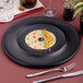 A large black round porcelain plate with a wide rim, displayed with a small gourmet food arrangement in the center.