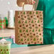 A natural kraft paper shopping bag with handles, decorated with a holiday print featuring Christmas trees, presents, candy canes, and holly.