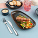 An oval asymmetric black porcelain coupe platter with grilled sausage and vegetables, accompanied by bread, sauce, and a drink.