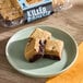 A plate with three square brookie brownie bites, featuring a cookie layer on top of a brownie layer, with a package of Killer Brownie Brookie Brownie Bites in the background.