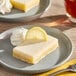 A rectangular lemon bar square garnished with a lemon slice and a dollop of whipped cream on a gray plate.