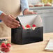 A black bakery box with a partially open lid containing cupcakes topped with strawberries.