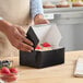 A black bakery box being closed by a person, containing desserts topped with strawberries.
