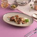 An oval beige porcelain coupe platter with a slightly raised rim, displayed on a pink surface and used for serving food.