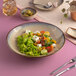 A beige round porcelain coupe deep plate filled with a fresh salad, displayed on a pink table.