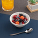 A small ivory round porcelain bowl filled with assorted berries and melon balls, accompanied by a spoon.