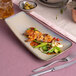 An asymmetric rectangular beige porcelain coupe platter with a food presentation, placed on a pink and beige table setting.