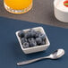 A small ivory square porcelain bowl filled with blueberries, placed next to a spoon.