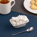 A small ivory rectangular porcelain bowl filled with sugar cubes, placed next to a spoon, a cup of tea, and a plate of cookies.