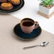 A blue porcelain espresso cup with a matching saucer, filled with coffee and placed on a beige placemat next to a spoon.
