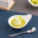 A small ivory porcelain teardrop-shaped bowl filled with a green sauce and a dumpling, placed next to a metal spoon.