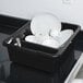 A black Tablecraft divided bus tub on a counter with white dishes and utensils.