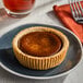 A small round pumpkin pie with a golden-brown filling and a thick, fluted crust, served on a dark plate.