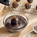 A bowl of melted dark chocolate compound coating being used to dip a strawberry, with finished chocolate-covered strawberries topped with sprinkles on a cooling rack in the background.
