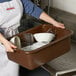 A woman in an apron holding a brown Tablecraft bus tub full of dishes.