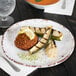 A white, irregular round melamine coupe plate with a rustic brown rim, holding a serving of grilled vegetables and a patty garnished with a lemon slice.