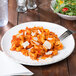 A white, round melamine coupe plate with an irregular edge, shown holding a serving of pasta with tomato sauce and cheese.